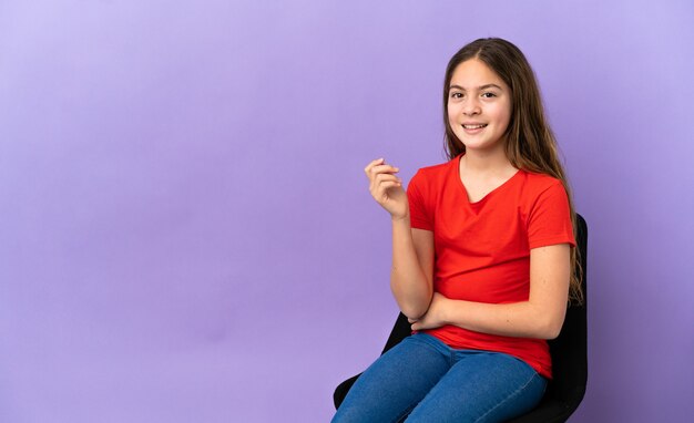 Kleines kaukasisches Mädchen, das auf einem Stuhl lokalisiert auf violettem Hintergrund sitzt und lacht
