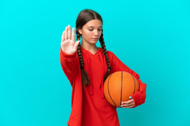 Kleines kaukasisches Mädchen, das Basketball einzeln auf blauem Hintergrund spielt, Stoppgeste macht und enttäuscht