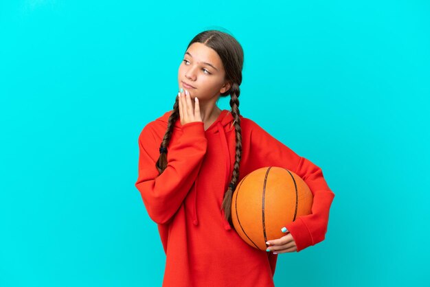 Kleines kaukasisches Mädchen, das Basketball isoliert auf blauem Hintergrund spielt und lächelnd nach oben schaut