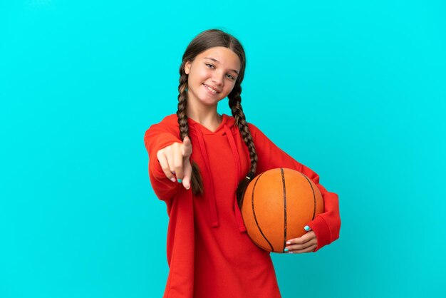 Kleines kaukasisches Mädchen, das Basketball isoliert auf blauem Hintergrund spielt und nach vorne mit glücklichem Ausdruck zeigt