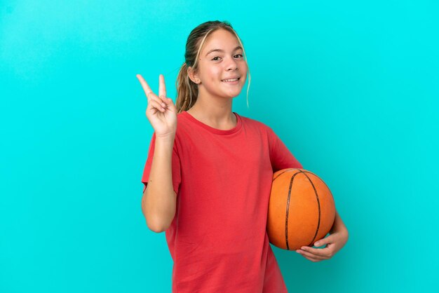 Foto kleines kaukasisches mädchen, das basketball spielt, isoliert auf blauem hintergrund, lächelt und zeigt victory-zeichen