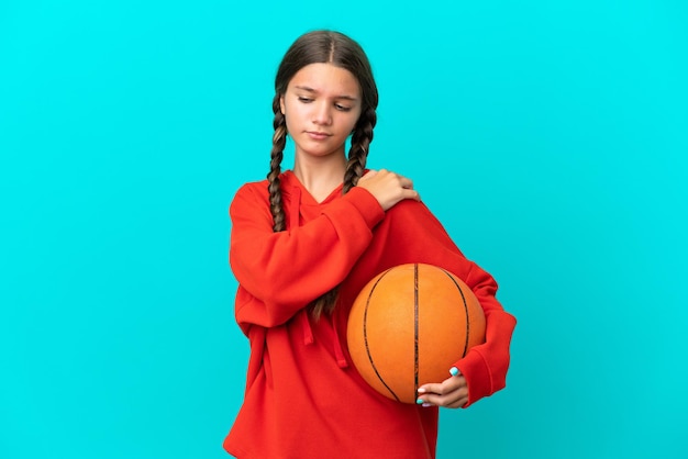 Kleines kaukasisches Mädchen, das Basketball spielt, isoliert auf blauem Hintergrund, leidet unter Schmerzen in der Schulter, weil es sich Mühe gegeben hat