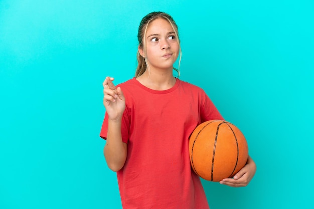 Kleines kaukasisches Mädchen, das Basketball spielt, isoliert auf blauem Hintergrund, mit gekreuzten Fingern und dem Wunsch nach dem Besten