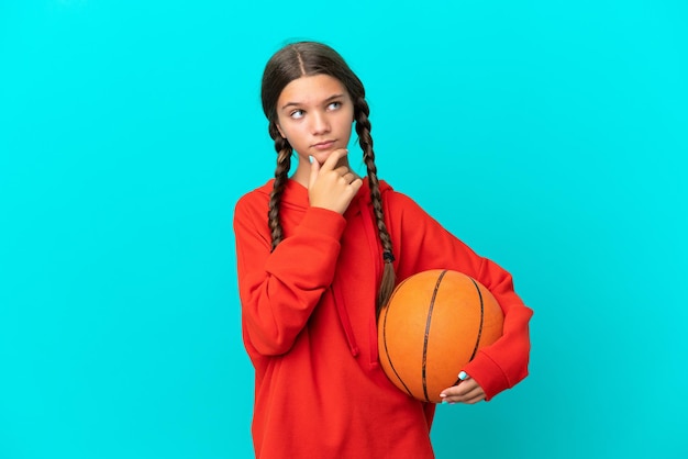 Kleines kaukasisches Mädchen, das Basketball spielt, isoliert auf blauem Hintergrund und Zweifel hat