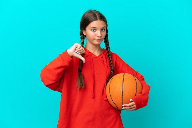Kleines kaukasisches Mädchen, das Basketball spielt, isoliert auf blauem Hintergrund, zeigt Daumen nach unten mit negativem Ausdruck