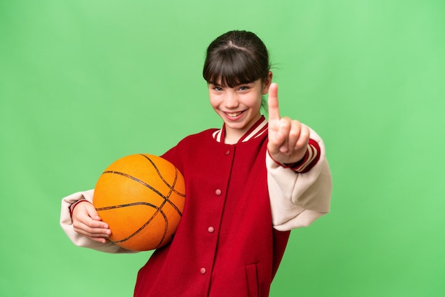 Kleines kaukasisches Mädchen, das Basketball vor isoliertem Hintergrund spielt und einen Finger zeigt und hebt