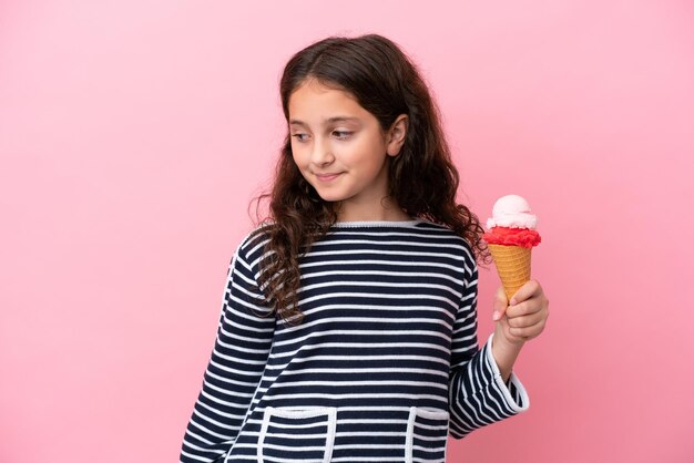 Kleines kaukasisches Mädchen, das ein Eis isoliert auf rosafarbenem Hintergrund hält, zur Seite schaut und lächelt