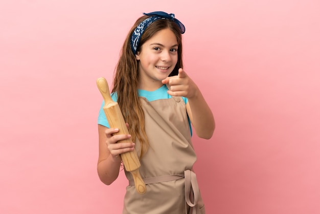 Kleines kaukasisches Mädchen, das ein Nudelholz isoliert auf rosa Hintergrund hält und nach vorne mit glücklichem Ausdruck zeigt