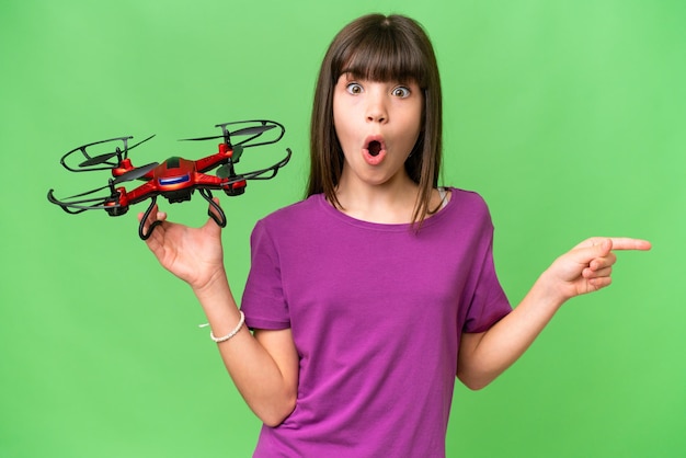 Kleines kaukasisches Mädchen, das eine Drohne über isoliertem Hintergrund hält, überrascht und zeigt zur Seite