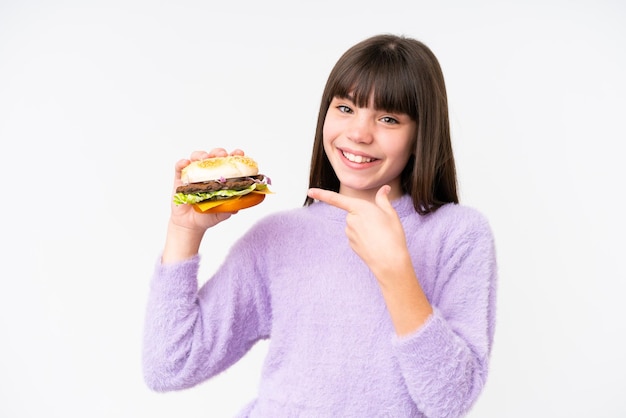 Kleines kaukasisches Mädchen, das einen Burger über isoliertem Hintergrund hält und darauf zeigt