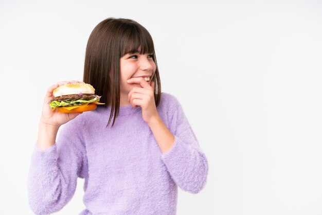 Kleines kaukasisches Mädchen, das einen Burger vor isoliertem Hintergrund hält, denkt an eine Idee und schaut zur Seite