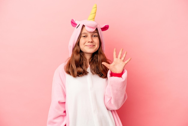Kleines kaukasisches Mädchen, das einen Einhorn-Pyjama trägt, isoliert auf rosafarbenem Hintergrund, lächelt fröhlich und zeigt Nummer fünf mit den Fingern.