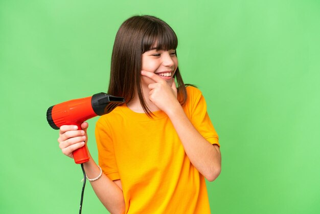 Kleines kaukasisches Mädchen, das einen Haartrockner über isoliertem Hintergrund hält und eine Idee denkt und zur Seite schaut