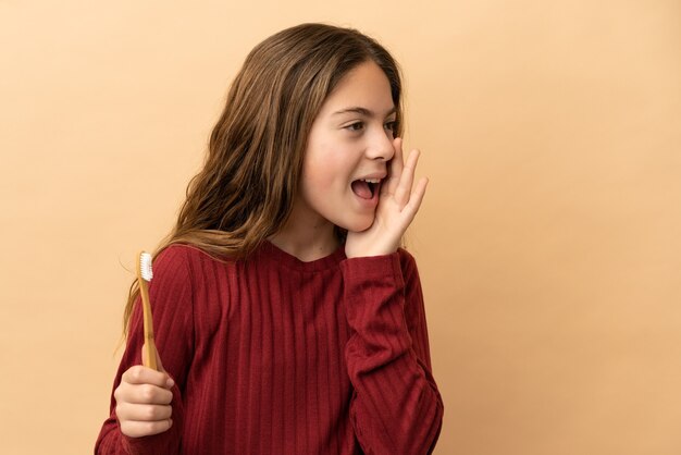 Kleines kaukasisches Mädchen, das sich die Zähne putzt, isoliert auf beigefarbenem Hintergrund, das mit weit geöffnetem Mund zur Seite schreit