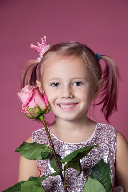 Kleines kaukasisches Mädchen in einem festlichen Kleid mit Pailletten, das mit rosafarbener Blume auf einem rosa Hintergrund in die Kamera schaut