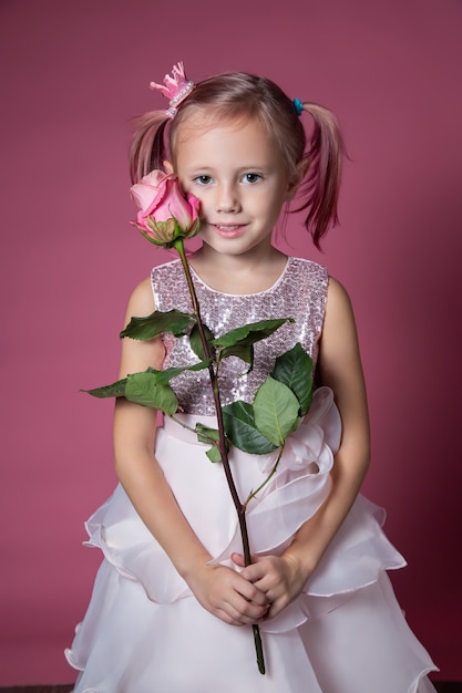 Kleines kaukasisches Mädchen in einem festlichen Kleid mit Pailletten, das mit rosafarbener Blume auf einem rosa Hintergrund in die Kamera schaut