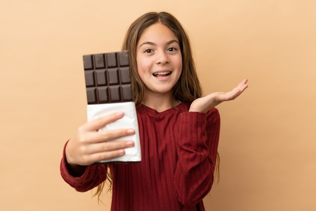 Kleines kaukasisches Mädchen isoliert auf beigefarbenem Hintergrund, das eine Schokoladentablette nimmt und überrascht