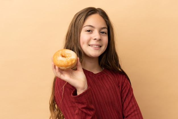 Kleines kaukasisches Mädchen isoliert auf beigefarbenem Hintergrund, das einen Donut hält und glücklich