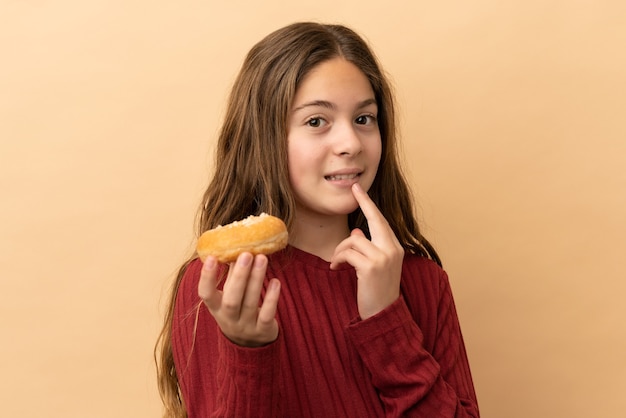 Kleines kaukasisches Mädchen isoliert auf beigefarbenem Hintergrund, das einen Donut hält