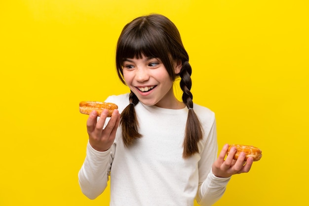 Kleines kaukasisches Mädchen isoliert auf gelbem Hintergrund, das Donuts mit glücklichem Ausdruck hält