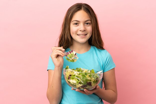 Kleines kaukasisches Mädchen isoliert auf rosa Hintergrund, das eine Schüssel Salat mit glücklichem Ausdruck hält