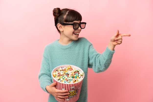 Kleines kaukasisches Mädchen lokalisiert auf Rosa mit 3D-Brille und hält einen großen Eimer Popcorn, während es nach vorne zeigt