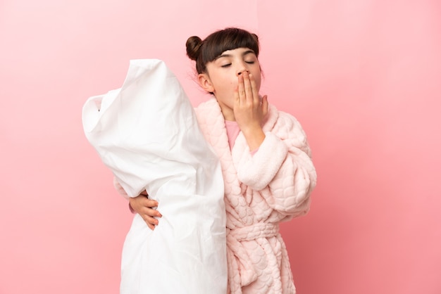 Kleines kaukasisches Mädchen lokalisiert auf rosa Wand im Schlafanzug und hält ein Kissen und Gähnen