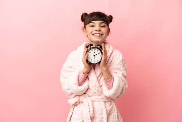 Kleines kaukasisches Mädchen lokalisiert auf rosa Wand im Schlafanzug und hält Uhr mit glücklichem Ausdruck