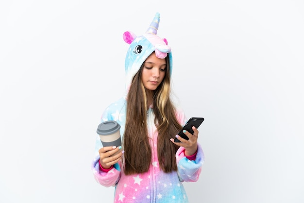 Kleines kaukasisches Mädchen mit Einhorn-Pyjama isoliert auf weißem Hintergrund mit Kaffee zum Mitnehmen und einem Handy