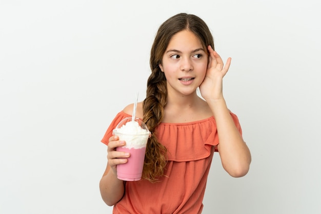 Kleines kaukasisches Mädchen mit Erdbeermilchshake isoliert auf weißem Hintergrund, das etwas hört, indem es die Hand auf das Ohr legt