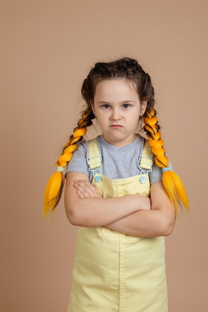 Kleines kaukasisches Mädchen mit gelben Kanekalon-Zöpfen, beleidigt von jemandem, der Wut mit verschränkten Armen in gelbem Overall und grauem T-Shirt auf beigem Hintergrund zeigt
