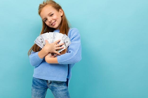 Kleines kaukasisches Mädchen mit langen Stirnhaaren im blauen Kapuzenpulli hält viel Geld isoliert auf Blau