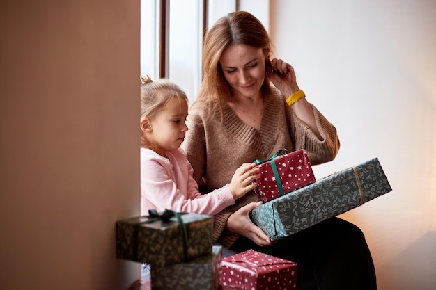 Kleines kaukasisches Mädchen mit Mutter, die zu Hause in der Nähe des Fensters Geschenkboxen auspackt Glückliches Familienweihnachtskonzept