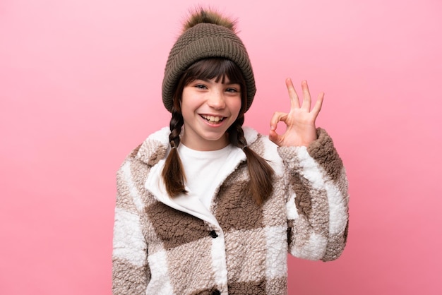 Kleines kaukasisches Mädchen mit Winterjacke isoliert auf rosa Hintergrund, das mit den Fingern ein Ok-Zeichen zeigt