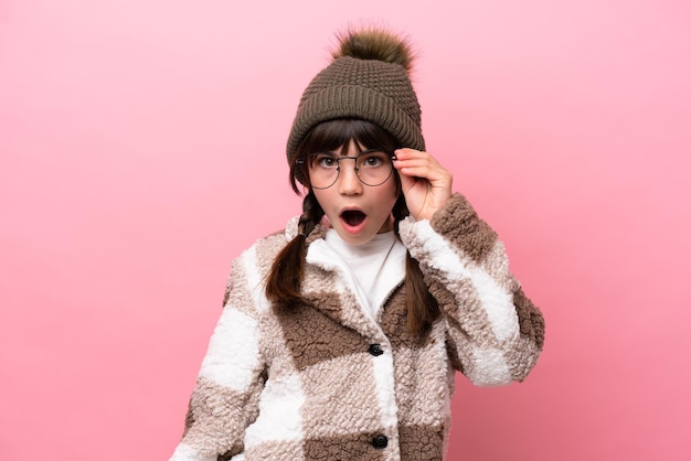 Kleines kaukasisches Mädchen mit Winterjacke isoliert auf rosa Hintergrund mit Brille und überrascht