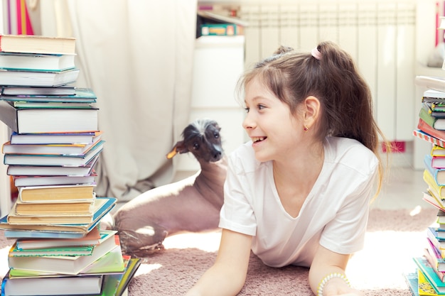 Kleines kaukasisches Mädchen spielt mit einem Hund auf dem Boden. Es gibt viele Bücher um sie herum. Das Mädchen versucht zu lesen, der Hund stört sie.