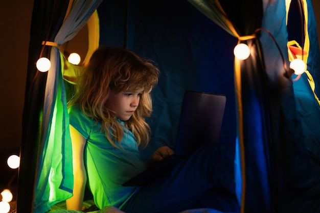 Kleines Kind benutzt nachts einen Laptop, ein Junge, der vom blauen Licht eines Computerbildschirms beleuchtet wird