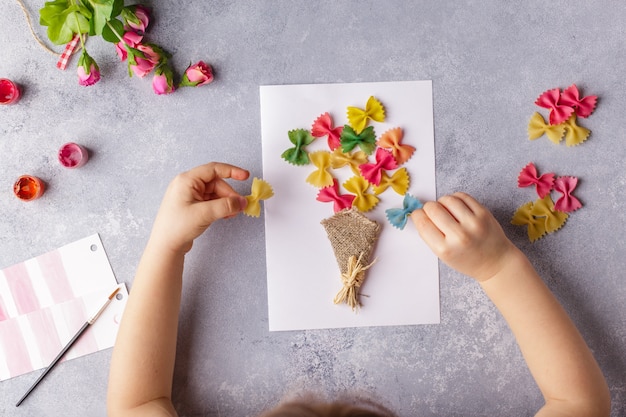 Kleines Kind, das einen Blumenstrauß aus farbigem Papier und farbigen Teigwaren heraus tut.