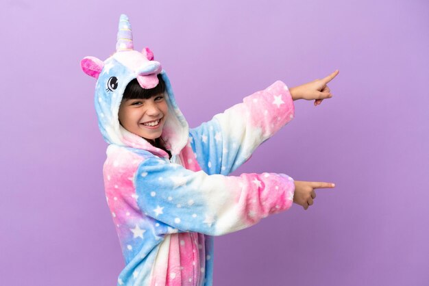 Kleines Kind, das einen Einhornpyjama trägt, isoliert auf violettem Hintergrund, zeigt mit dem Finger zur Seite und präsentiert ein Produkt