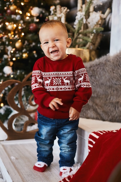 Kleines Kind im roten Weihnachtspullover posiert auf der Treppe des für Weihnachten dekorierten Hauses.