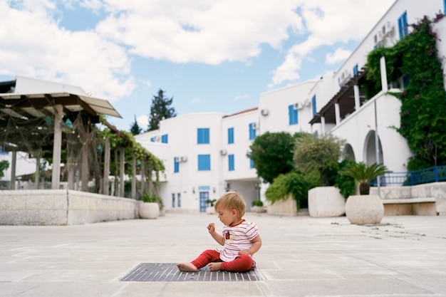 Kleines Kind sitzt auf einem Kanalgitter vor dem Hintergrund von Häusern und Pflanzen in Wannen