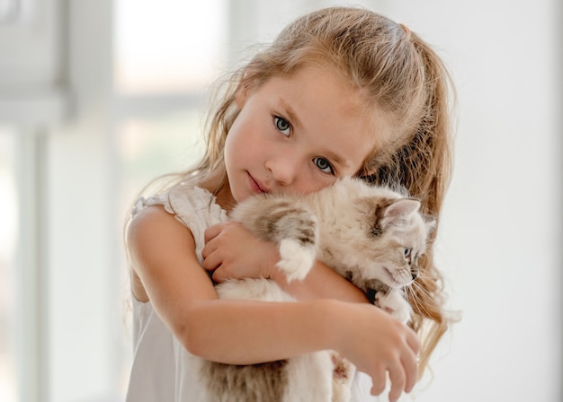 Kleines Kindermädchen, das Ragdollkätzchen hält und es umarmt. Hübsches weibliches Kind mit katzenartigem Hauskatzenhaustier zu Hause