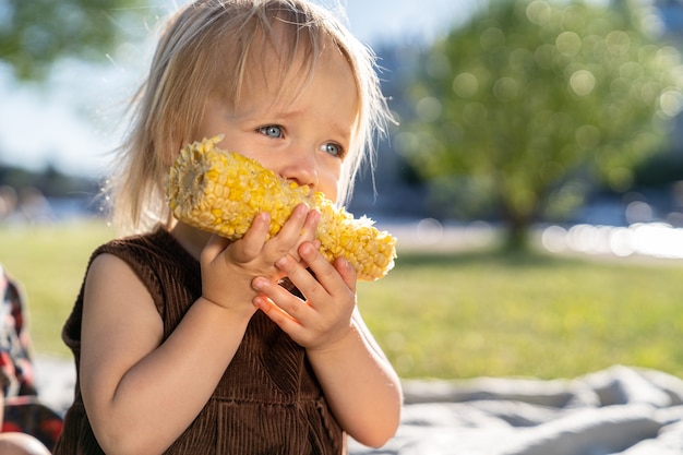 Kleines Kindermädchen essen Zuckermais, sitzend auf Plaid auf Gras im Sommertag. Gesundes Essen.