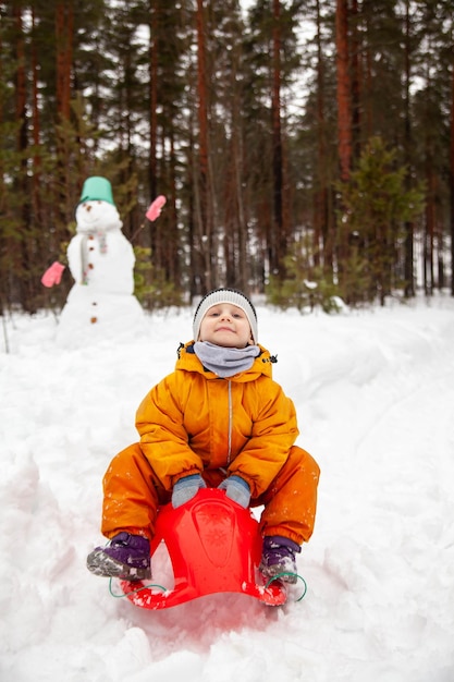 Kleines Kleinkind auf Winterspaziergang in der Nähe von Schneemann auf Schlitten