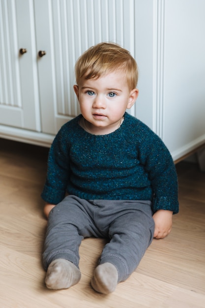 Kleines Kleinkind mit ansprechenden blauen Augen, prallen Wangen und blonden Haaren sitzt auf dem Boden