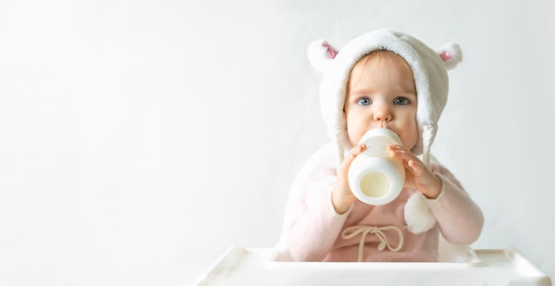 Kleines Kleinkindmädchen in einem warmen flauschigen Hut trinkt Milch von einer Flasche, während sie sitzt. grauer Hintergrund