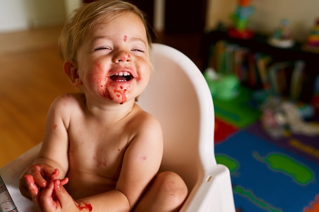 Kleines lachendes Baby mit schmutzigem Gesicht sitzt auf einem Stuhl mit verschränkten Armen