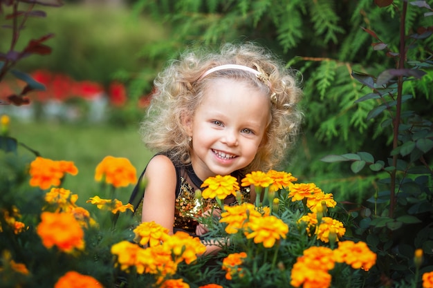 Kleines lachendes Mädchen mit lockigen blonden Haaren spielt mit orangefarbenen Blumen im Sommerpark