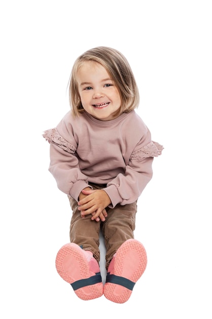 Kleines lachendes Mädchen sitzt auf dem Boden. Ein Kind in braunen Jeans und einem rosa Pullover. Liebe und Zärtlichkeit. Isoliert auf weißem Hintergrund. Vertikal.