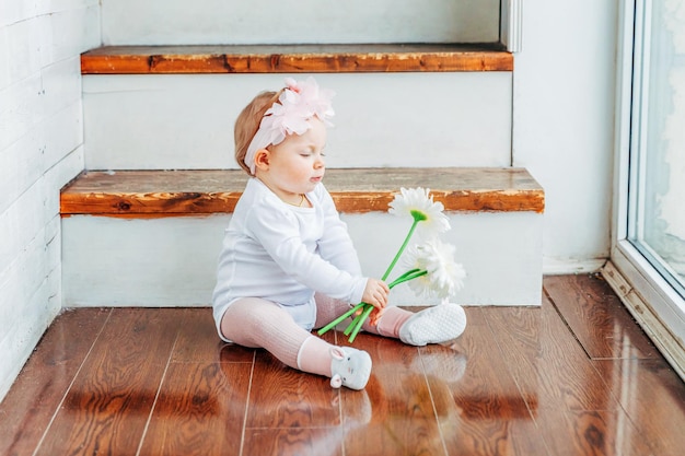 Kleines lächelndes Babymädchen, das ein Jahr alt ist und einen Frühlingskranz trägt, der sich auf dem Boden in einem hellen Wohnzimmer in der Nähe des Fensters befindet und mit Gerbera-Blumen spielt Glückliches Kind, das zu Hause spielt Kindheitskonzept